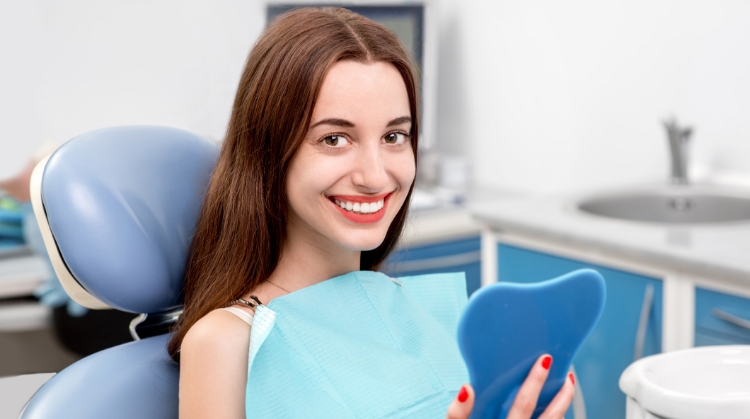 A smiling woman sitting in a dental chair, holding a blue dental mirror. She is wearing a dental bib and appears satisfied with her dental care in a clean, modern dental office.