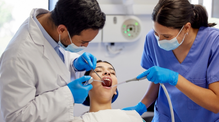 A dentist and a dental assistant performing a dental cleaning on a patient in a clinic. The patient is reclining in the chair with their mouth open while the dentist uses dental instruments, and the assistant provides suction.